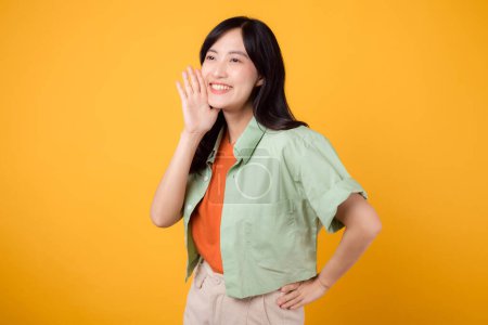 Energetic young 30s Asian woman wearing a green shirt on an orange background, enthusiastically shouting with excitement. Explore the concept of discount shopping promotion with this vibrant image.