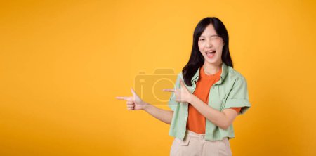 Téléchargez les photos : Jeune femme asiatique trentaine portant une chemise verte sur une chemise orange, pointant du doigt pour libérer de l'espace de copie. Découvrez le monde passionnant de la promotion des achats discount - en image libre de droit