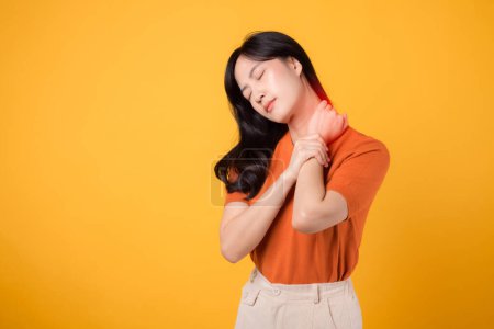 Téléchargez les photos : Femme asiatique préoccupée trentaine, portant une chemise orange, tient son cou douloureux sur fond jaune. syndrome de bureau concept de soins de santé. - en image libre de droit