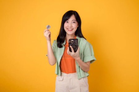 Téléchargez les photos : Jeune femme asiatique 30s visage heureux habillé en chemise orange et pullover vert montrant pièce de monnaie crypto tout en tenant smartphone isolé sur fond jaune. Concept de financement futur. - en image libre de droit