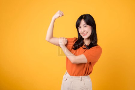 Téléchargez les photos : Célébrez avec confiance comme une jeune femme asiatique trentaine présente un geste de signe de la main poing vers le haut, portant une chemise orange sur fond jaune. Concept d'autonomisation et de féminisme. - en image libre de droit