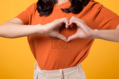 Foto de Salud, bienestar y bienestar con una joven mujer asiática de 30 años. Muestra un gesto de la mano del corazón en su pecho, con una camisa naranja, sobre un fondo amarillo vibrante. - Imagen libre de derechos