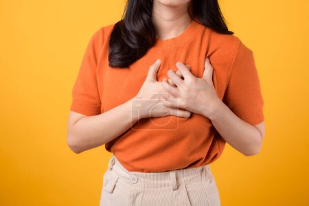 Téléchargez les photos : Gros plan de la femme asiatique de soutien trentaine, portant une chemise orange, tient la main sur la poitrine sur fond jaune. crise cardiaque maladie, douleur thoracique concept de soins de santé. - en image libre de droit
