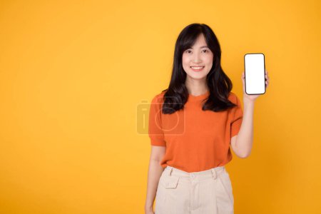 Photo for Young 30s asian woman wearing orange shirt showing smartphone blank display screen isolated on yellow background. cheerful happy face female person with new application technology concept. - Royalty Free Image