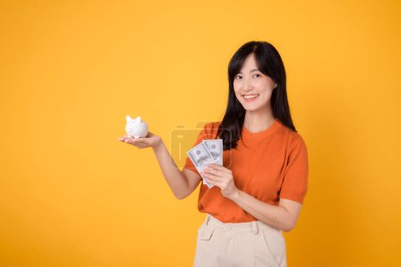 Photo for Joyful Asian woman 30s holding piggy bank and cash money dollars on vibrant yellow background. Saving money concept. - Royalty Free Image