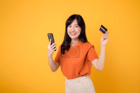 Energetische junge Asiatin um die 30, mit Smartphone und Kreditkarte auf gelbem Hintergrund. Schnelles Online-Shopping.