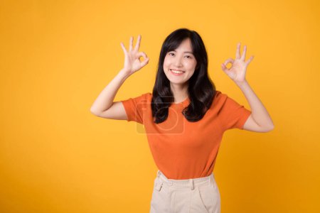 Photo for A young Asian woman 30s, wearing an orange shirt, showcases the okay sign gesture on a sunny yellow background. Hands gesture concept. - Royalty Free Image