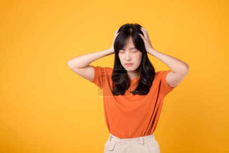 Photo for Expressive portrait of young asian woman 30s in distress, standing in a yellow studio background, illustrating stress, headache, and health concerns. - Royalty Free Image