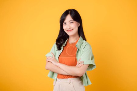 Photo for Beauty of confidence and well-being through young 30s Asian woman wearing orange shirt exudes grace with arm cross gesture on chest against yellow background, captivating portrait of self-assurance. - Royalty Free Image