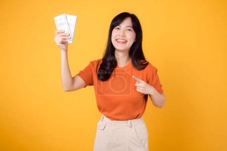 Foto de Mujer asiática enérgica 30s puntos a dinero en efectivo dólares en vibrante fondo amarillo. Concepto de libertad financiera. - Imagen libre de derechos