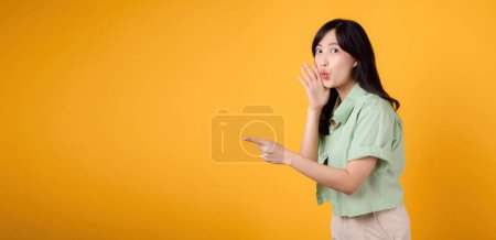 Téléchargez les photos : Capture fiançailles et l'excitation avec la jeune femme asiatique porte une chemise vert pastel sur chemise orange montrent sourire heureux tout en plaçant la main sur la bouche et pointant avec un doigt pour libérer de l'espace de copie. - en image libre de droit