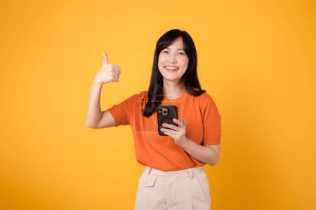 Foto de Mujer asiática joven y enérgica de 30 años, con camisa naranja, usando un teléfono inteligente con un cartel de mano con el pulgar hacia arriba sobre un vibrante fondo amarillo. Nuevo concepto de aplicación móvil. - Imagen libre de derechos