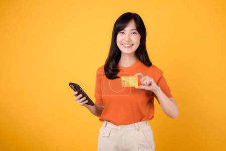 Vivid Asian woman 30s, utilizing smartphone and showcasing credit card on vibrant yellow background. Quick online shopping payment process.