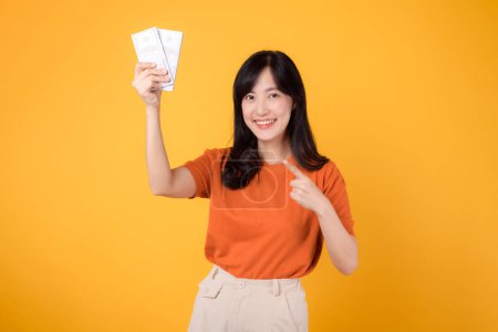 Photo for Dynamic Asian woman 30s points to cash money dollars on vibrant yellow backdrop. Wealth and prosperity concept. - Royalty Free Image