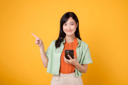 young asian woman 30s happy smile dressed in orange shirt showing smartphone with pointing finger hand gesture to free space isolated on yellow studio background. app smartphone concept