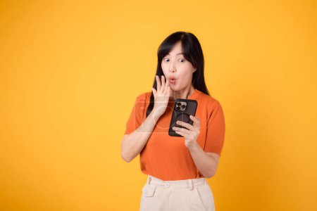 Photo for Excited Asian woman 30s, wearing orange shirt, using smartphone with fist up hand sign on vibrant yellow background. Thrilling new app experience. - Royalty Free Image