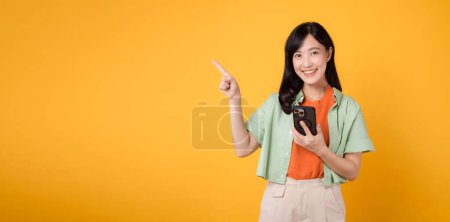 Mobile Apps mit einer fröhlichen jungen Asiatin in den 30er Jahren, die ein orangefarbenes Hemd trägt, zeigt fröhlich auf ihr Smartphone und zeigt mit der Fingerbewegung isoliert auf gelben Studiohintergrund. Smartphone-App-Konzept.