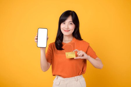 Photo for Joyful Asian woman 30s, revealing blank smartphone screen and credit card on vibrant yellow background. Swift online payment shopping. - Royalty Free Image