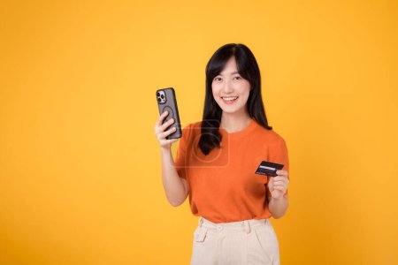 Téléchargez les photos : Jeune femme asiatique excitée dans la trentaine en chemise orange, utilisant un smartphone et tenant une carte de crédit sur fond jaune. Délicieux achats en ligne. - en image libre de droit