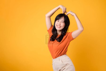 Téléchargez les photos : Rayonner de positivité et de connexion comme une femme asiatique des années 30 porte une chemise orange, formant une forme de coeur avec ses bras, symbolisant l'amour et l'unité. - en image libre de droit