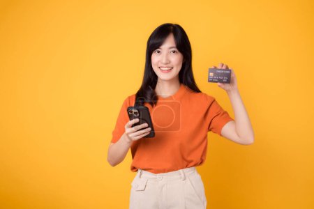 Foto de Mujer asiática joven y enérgica de 30 años, usando un teléfono inteligente y sosteniendo la tarjeta de crédito sobre un fondo amarillo. Compras en línea sin fisuras. - Imagen libre de derechos