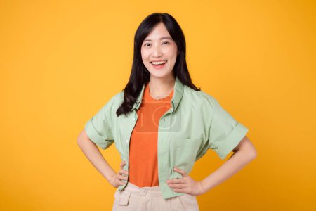 Téléchargez les photos : Bonheur rayonnant jeune femme asiatique trentaine portant une chemise verte sur une chemise orange met en valeur le visage joyeux tout en se tenant debout avec les bras akimbo. pure joie avec une image vibrante sur un fond jaune. - en image libre de droit