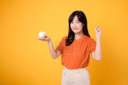 Photo for Joyful Asian woman 30s holding piggy bank, showing zero hand sign, on vibrant yellow background. Wealth saving concept. - Royalty Free Image