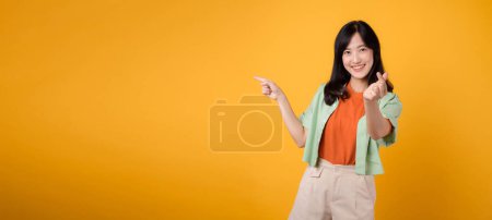 Foto de Joven mujer asiática de 30 años con una camisa verde sobre un fondo naranja señala los dedos a espacio libre copia mientras que muestra un mini signo de la mano del corazón. Explora el mundo del descuento concepto de promoción de compras. - Imagen libre de derechos