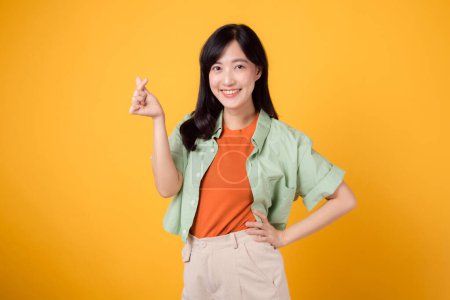 Foto de Felicidad con una joven mujer asiática de 30 años, vestida con una camisa naranja y un jersey verde. Su mini gesto del corazón, la sujeción de la cadera y su suave sonrisa transmiten un mensaje profundo a través del lenguaje corporal. - Imagen libre de derechos