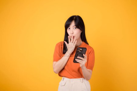Photo for Intrigued Asian woman 30s, wearing orange shirt, using smartphone with fist up hand sign on vibrant yellow background. Fascinating new app exploration. - Royalty Free Image