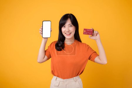 Téléchargez les photos : Femme asiatique dynamique 30s, révélant smartphone et carte de crédit sur fond jaune. Achats efficaces de paiement en ligne. - en image libre de droit