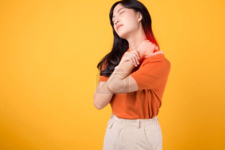 Foto de Mujer asiática vigilante de 30 años, vistiendo una camisa naranja, sostiene su doloroso cuello sobre un fondo amarillo. síndrome de oficina concepto de atención médica. - Imagen libre de derechos