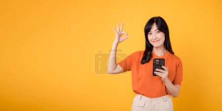 Photo for Vibrant Asian woman 30s, wearing orange shirt, using smartphone with okay hand sign on vibrant yellow background. New mobile app concept. - Royalty Free Image