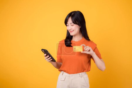 Youthful Asian woman 30s, using smartphone and displaying credit card on vibrant yellow background. Rapid online shopping payment experience.
