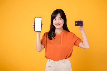 Photo for Vibrant Asian woman 30s, holding smartphone and credit card on yellow background. Hassle-free online payment shopping. - Royalty Free Image