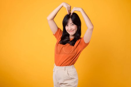 Foto de Ilustre el romance y las emociones sentidas con una mujer asiática de 30 años usando una camisa naranja, mostrando una sonrisa alegre y un gesto en forma de corazón. - Imagen libre de derechos