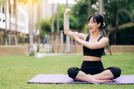Téléchargez les photos : Portrait fit belle jeune femme asiatique portant des vêtements de sport noirs écouter de la musique relax et montrant un geste de santé fort à son bras tout en faisant du yoga sur tapis de yoga dans le parc extérieur. - en image libre de droit