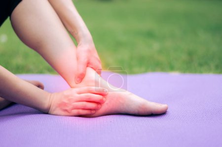 Téléchargez les photos : Sportive tenant sa cheville de douleur après avoir fait de l'exercice sur tapis de yoga dans le parc extérieur. accident de blessure dans le concept d'exercice. - en image libre de droit