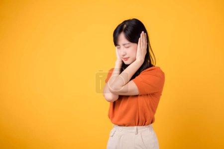 Téléchargez les photos : Femme asiatique fatiguée criant en réaction à un bruit fort, couvrant son oreille. Expression de stress et de frustration isolée sur fond jaune. - en image libre de droit