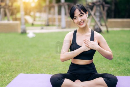 Foto de Retrato en forma delgada mujer asiática joven de 30 años que usa ropa deportiva negra cogida de la mano en el pecho mientras escucha música relajante y se sienta en la esterilla de yoga en el parque público. Concepto de salud y bienestar. - Imagen libre de derechos