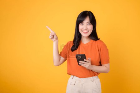 Photo for Smiling Asian woman 30s, wearing orange shirt, using smartphone, pointing to free copy space on vibrant yellow background. New mobile app concept. - Royalty Free Image