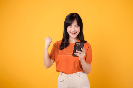 Foto de Mujer asiática alegre de 30 años, vistiendo una camisa naranja, usando un teléfono inteligente con el puño hacia arriba letrero de mano sobre un vibrante fondo amarillo. Celebra el nuevo concepto de aplicación. - Imagen libre de derechos