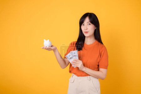 Photo for Happy Asian woman 30s holding piggy bank and cash money dollars on vibrant yellow background. Financial wealth concept. - Royalty Free Image