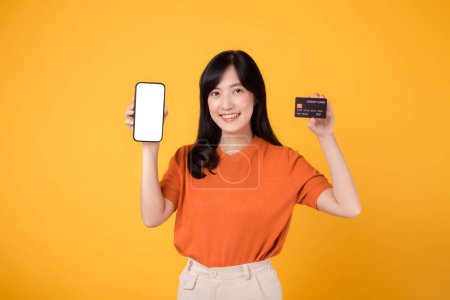 Excited Asian woman 30s, presenting smartphone and credit card on yellow background. Convenient online payment shopping.