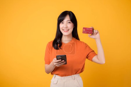 Foto de Mujer asiática emocionada en sus 30 años en camisa naranja, usando un teléfono inteligente y mostrando la tarjeta de crédito en el fondo amarillo. Compras rápidas en línea. - Imagen libre de derechos