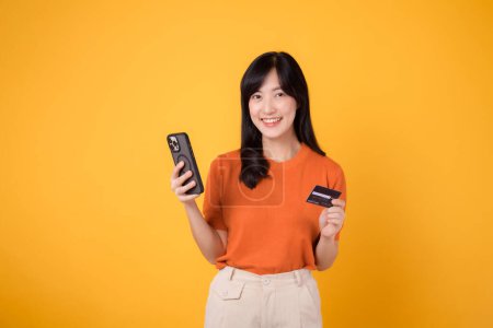 Photo for Excited young Asian woman in her 30s in orange shirt, using smartphone and holding credit card on yellow background. Enjoyable online shopping. - Royalty Free Image