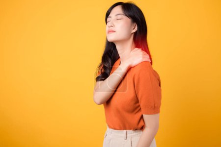 Photo for Neck ache therapy medical office syndrome concept. Concerned young Asian woman 30s, wearing an orange shirt, holds her pain shoulder on yellow background. - Royalty Free Image