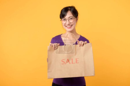 Téléchargez les photos : Jolie joyeux attrayant asiatique jeune femme portant chemise violette avec le visage de bonheur tenant vente sac de papier commercial isolé sur fond jaune. Concept de femme Shopaholic. - en image libre de droit