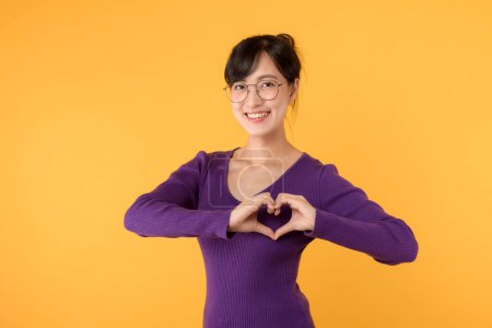 Photo for Portrait cheerful young Asian woman happy smile wearing purple sweater shirt expressing heart hand sign gesture on chest isolated yellow background. Valentine romantic symbol advertising concept. - Royalty Free Image