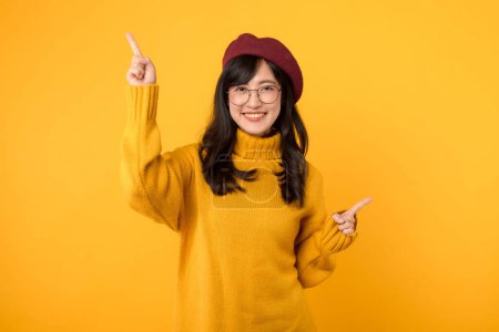 Téléchargez les photos : Portrait joyeux jeune femme asiatique portant un pull jaune et chapeau rouge exprimant les doigts pointant vers l'espace libre de copie pour la publicité isolée sur fond jaune. - en image libre de droit